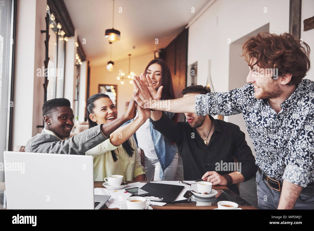 Happy young entrepreneurs in casual clothes at cafe table or in business office giving high fives to each other as if celebrating success or starting new project Stock Photo