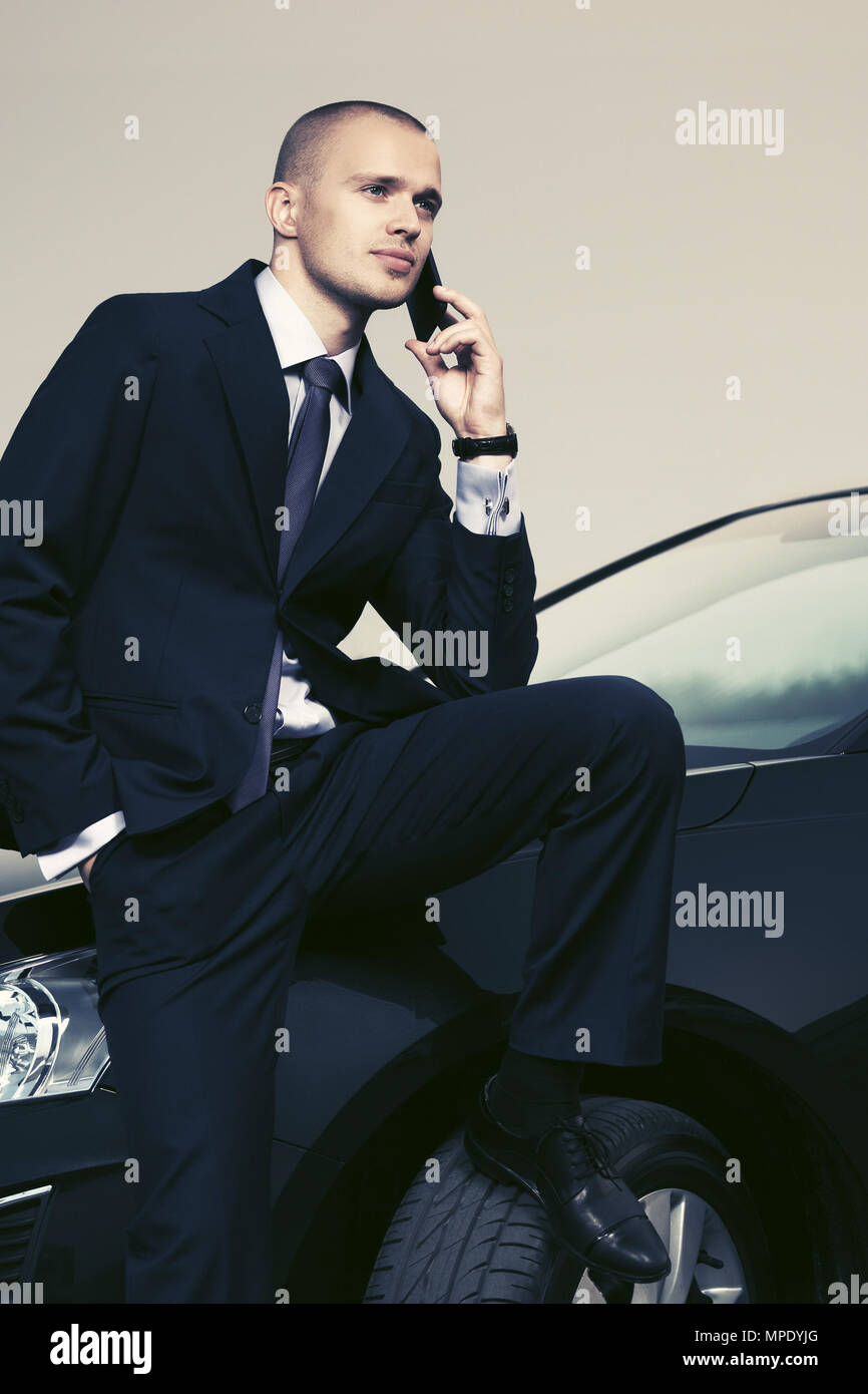 Young businessman in dark blue suit calling on cell phone sitting on his car Stock Photo