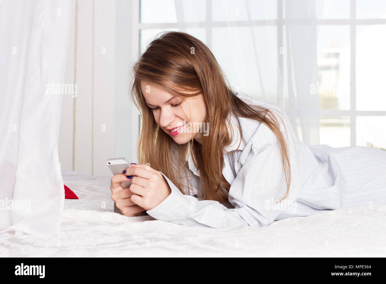 girl lies in a man's shirt on the bed with a smartphone in the hands of Stock Photo