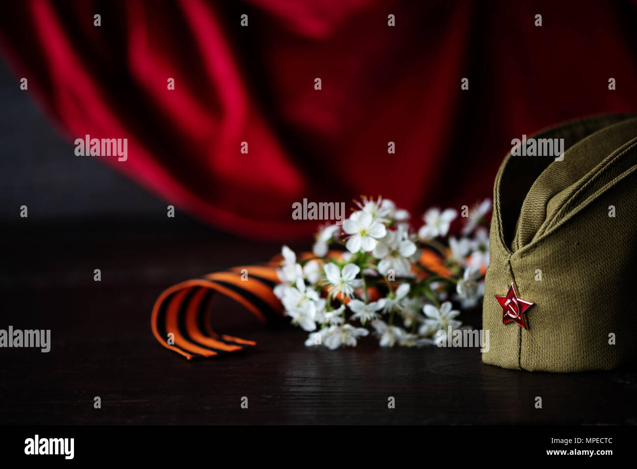 Concept, greeting card to the Victory Day. Soldier's cap with red star, cherry blossom and guards ribbon on the background of the red standard Stock Photo