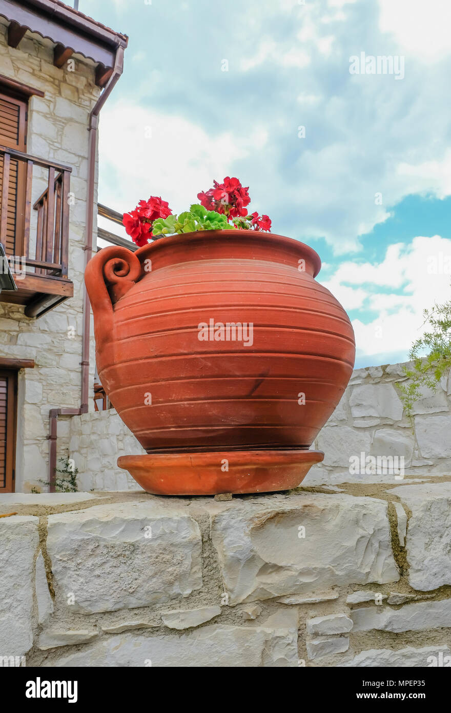 Round terracotta plant pot up on top of a stone wall with a red geranium plant flowering in early summer. Stock Photo