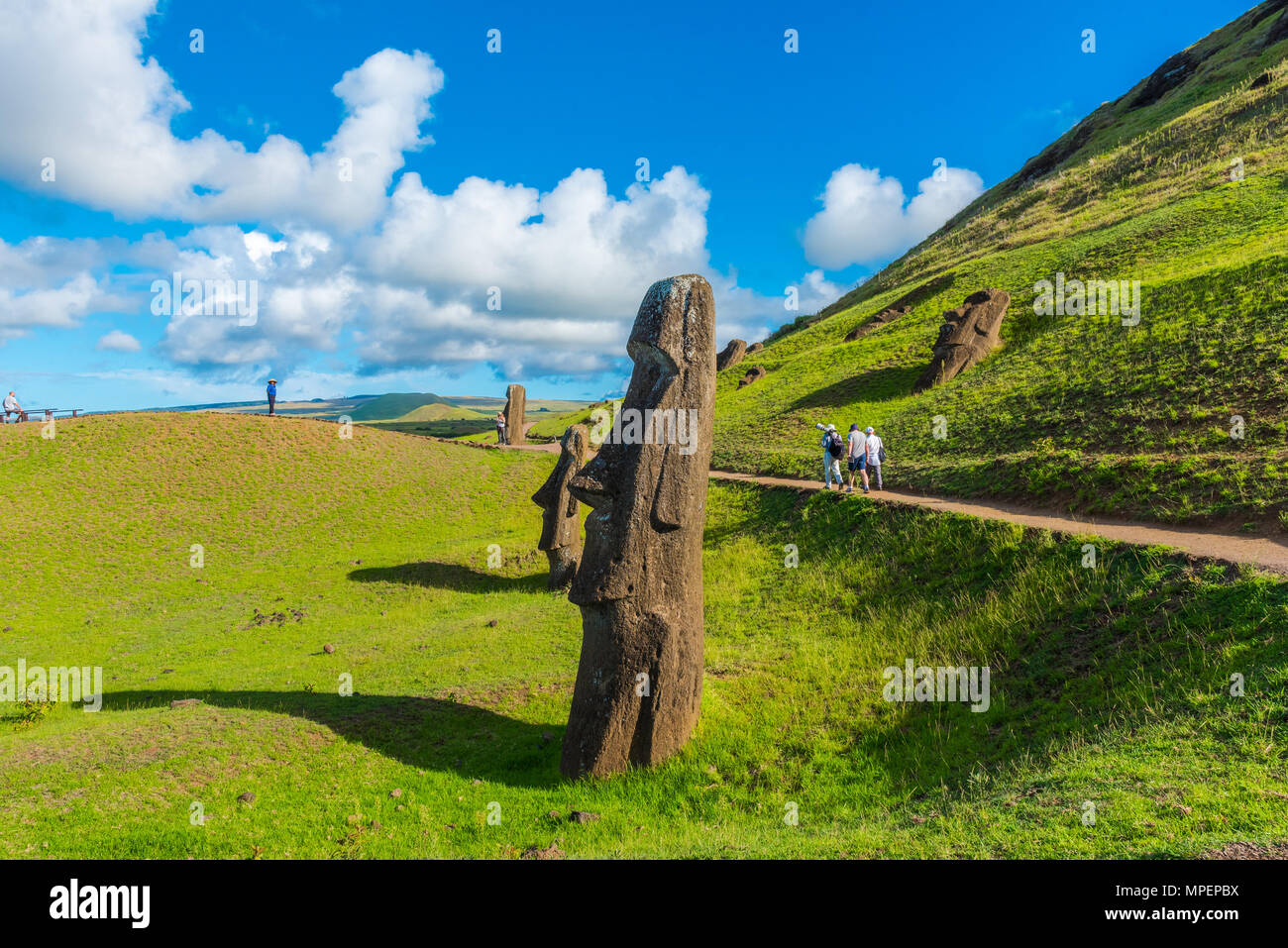 Moais line a road by a hill; in the distance tourists are walking by. Stock Photo