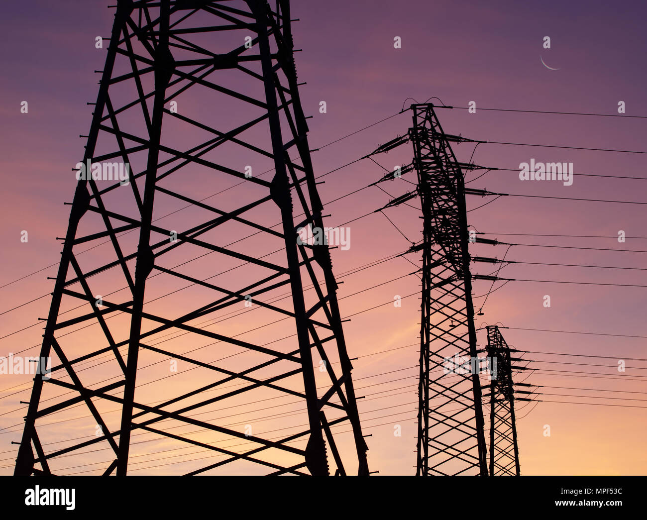 Three electricity pylons with cables are staggered as silhouettes in front of a colored evening sky. The moon can be seen as a narrow sickle. Stock Photo