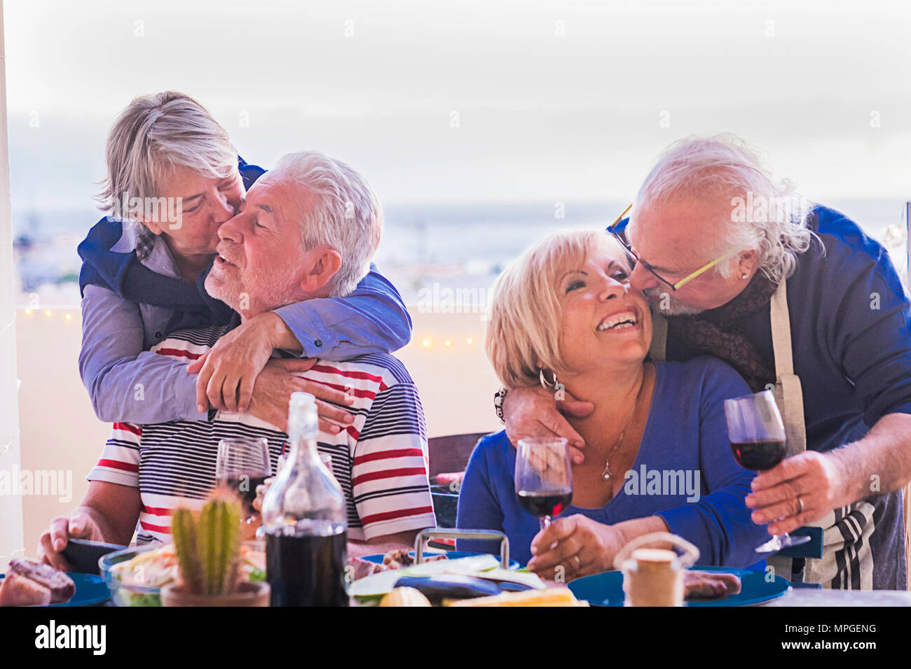 4 elderly gentlemen with gray hair hug and kiss while sitting at the table on the terrace. In the background yellow lights and ocean. With smiles and  Stock Photo