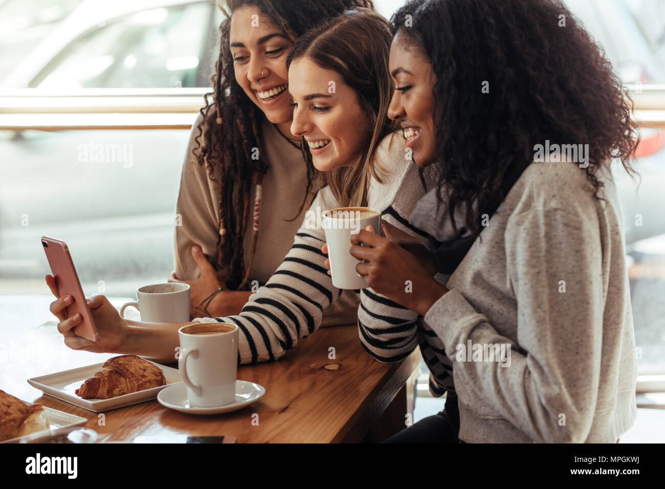 Three women sitting in a restaurant looking at mobile phone and smiling. Friends sitting at a cafe with coffee and snacks on the table looking at a mo Stock Photo