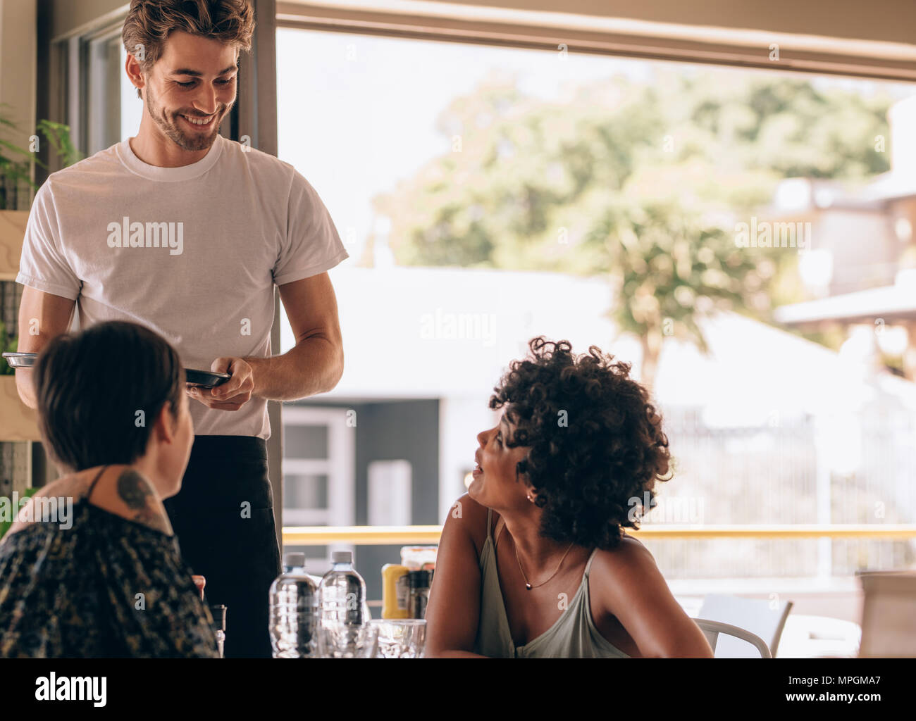 Young woman with a friend ordering to waiter at cafe. Two women sitting at cafe giving order to male waiter. Stock Photo