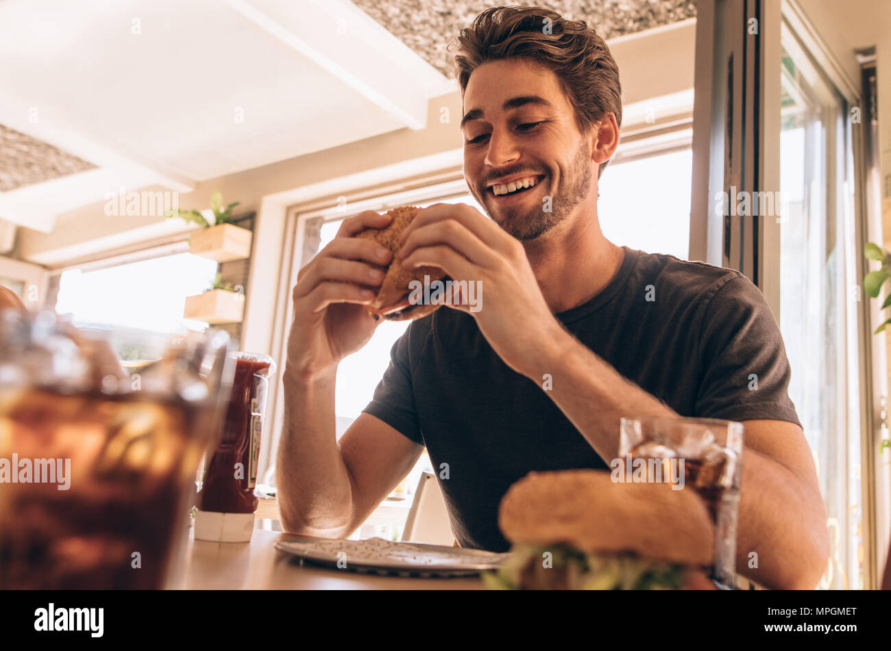 Happy young man eating a delicious burger at restaurant. Hungry young guy having stacked burger at fast food cafe. Stock Photo