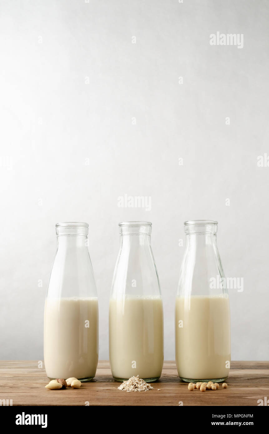A row of three glass bottles containing dairy free milk alternatives. Key ingredient in foreground on wood planked table. Almond, oat and soya varieti Stock Photo