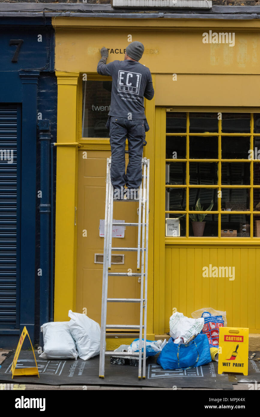 a man using a ladder to paint a building yellow. Man up a ladder on the
