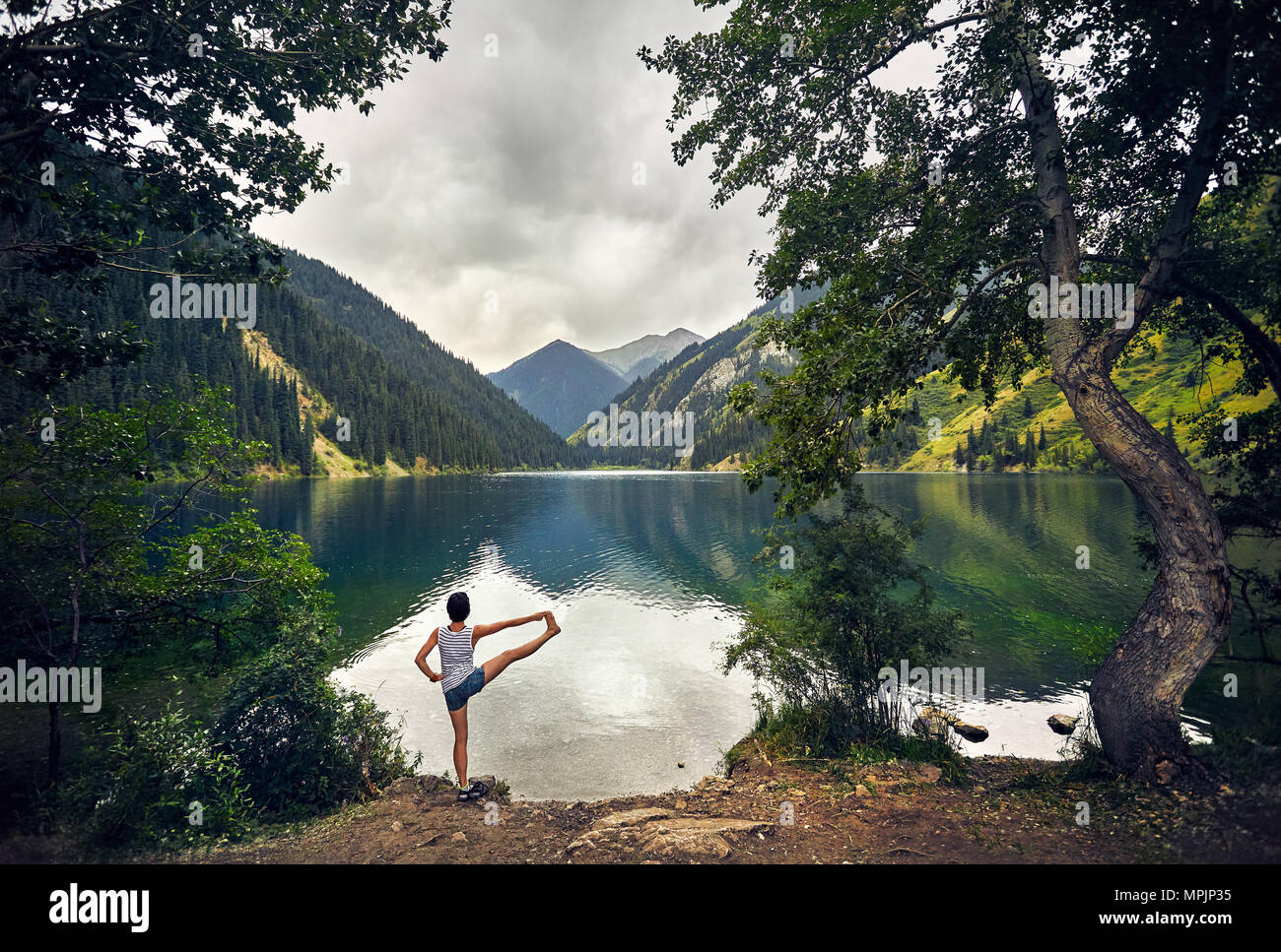 Young woman is practicing yoga balance pose at mountain lake with overcast sky background Stock Photo