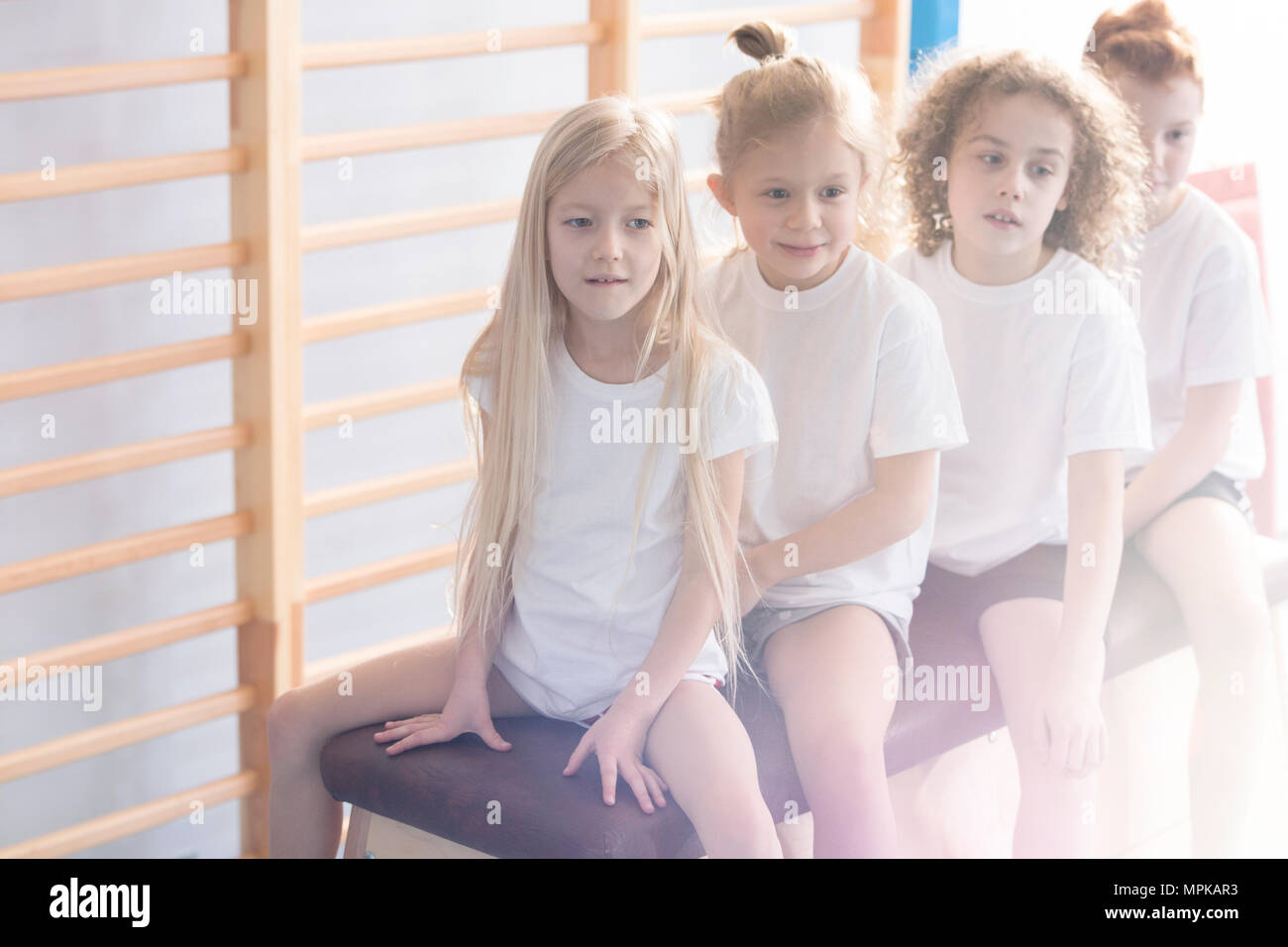 Close-up of a group of young children in white t-shirts sitting on a vaulting box looking in the direction of physical education teacher Stock Photo