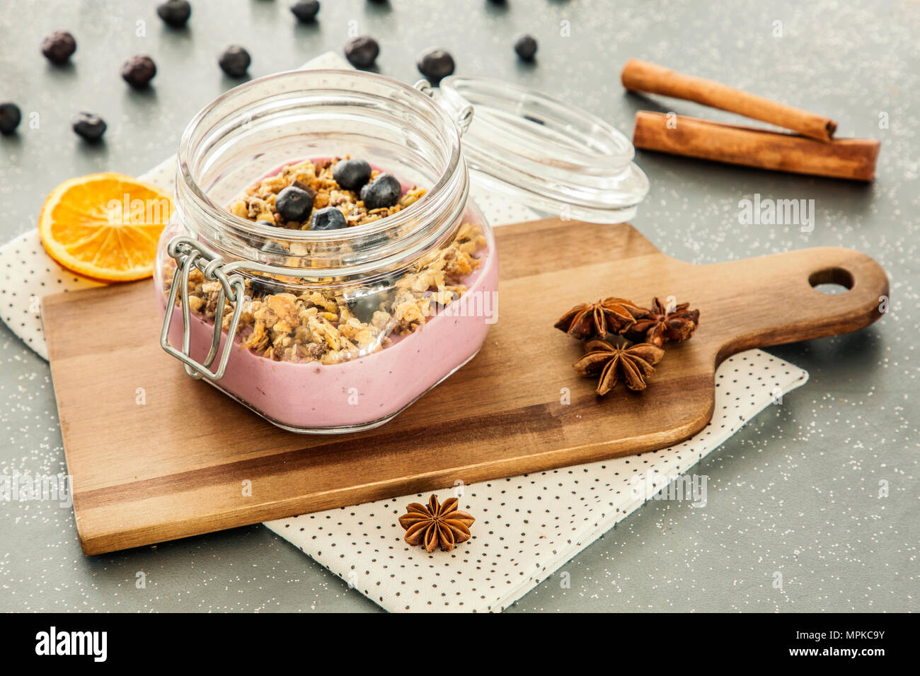Pink, fruit yogurt with granola and blueberries in a jar on a wooden cutting board and cinnamon spice on a gray kitchen counter Stock Photo
