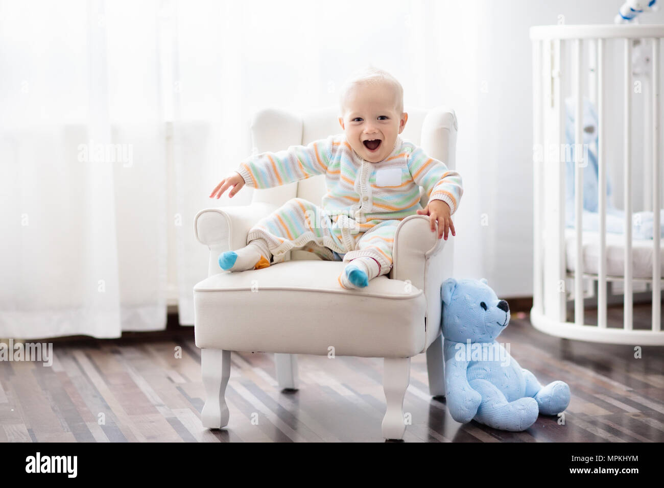 Baby boy in bedroom. Infant child sitting in white chair at round wooden bed. Kids room interior ...