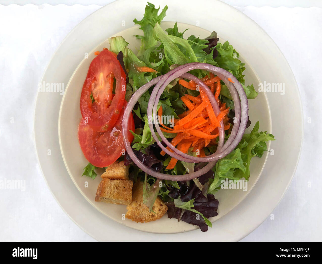 Small lettuce salad on a white plate, grated carrots, onions, tomato and croutons on a white placemat Stock Photo