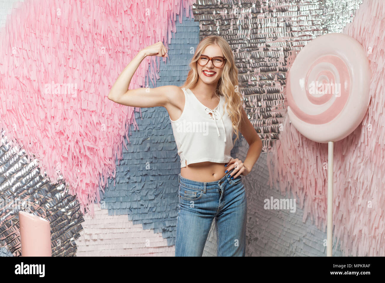 Funny young blonde girl with long hair in red glasses shows her muscles on colorful abstract background with huge candy, dressed in white top and jean Stock Photo