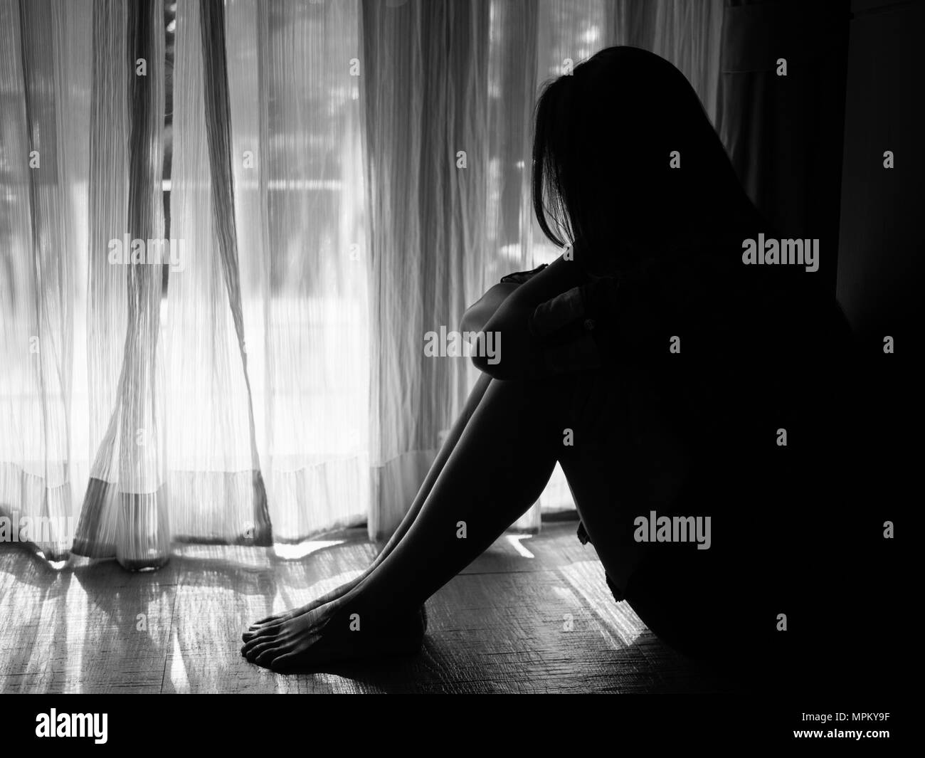 black and white of sad woman hug her knee and cry. Sad woman sitting alone in a empty room beside window or door Stock Photo