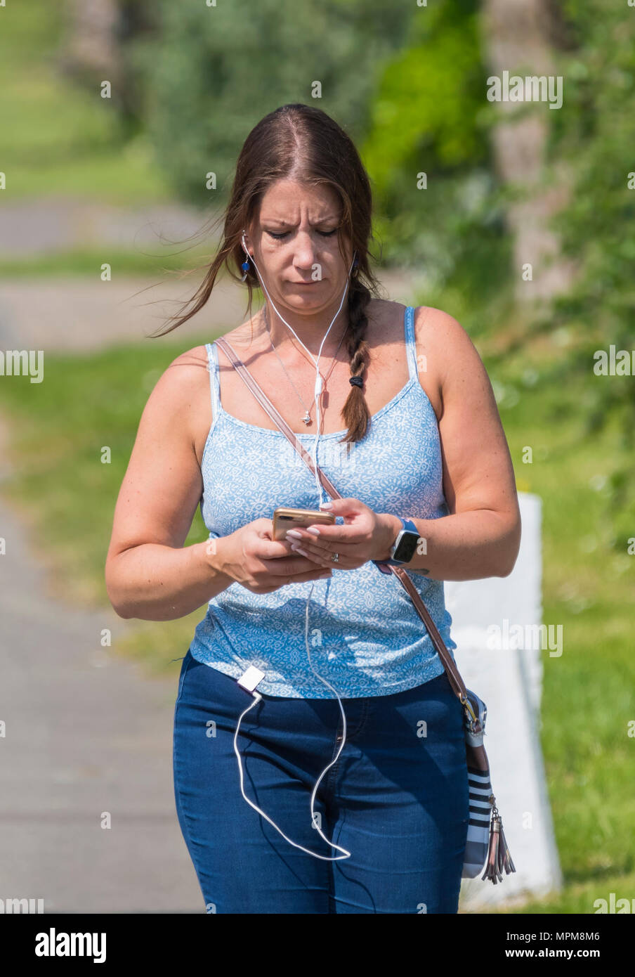 Young woman walking while looking down at a phone and also listening to music with in-ear headphones, on a hot summer day in the UK. Front facing. Stock Photo