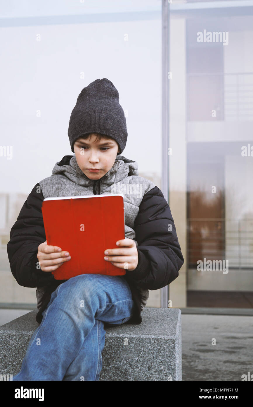 7 year old boy reading on tablet pc computer while sitting outside alone on city street in winter Stock Photo