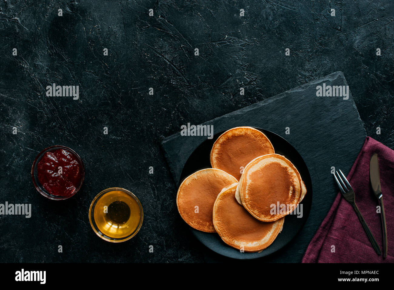top view of pancakes with jam and maple syrup on black concrete surface