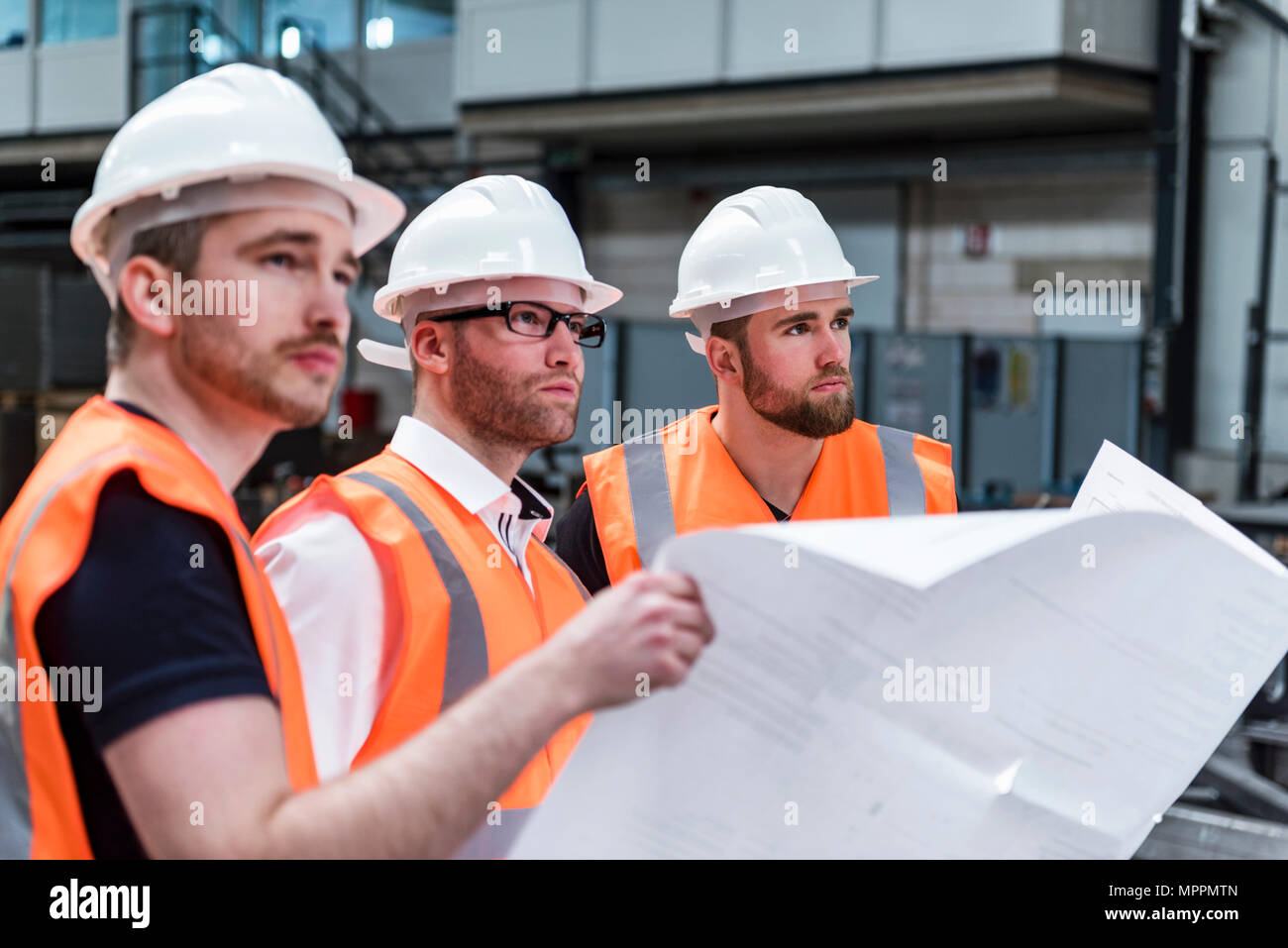 Three men wearing hard hats and safety vests holding plan in factory Stock Photo