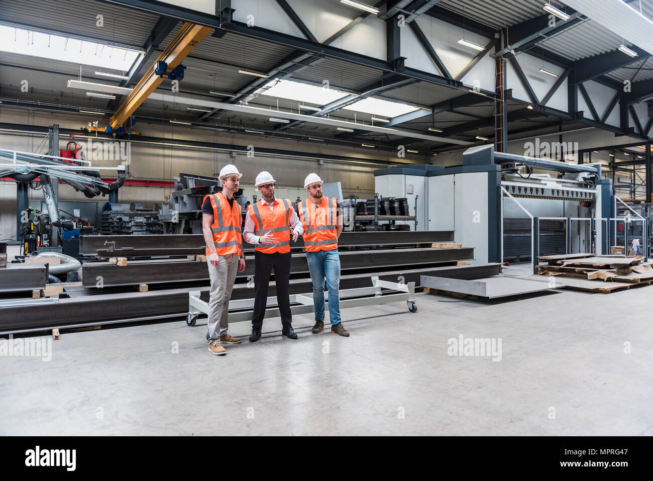 Three men wearing hard hats and safety vests talking on factory shop floor Stock Photo