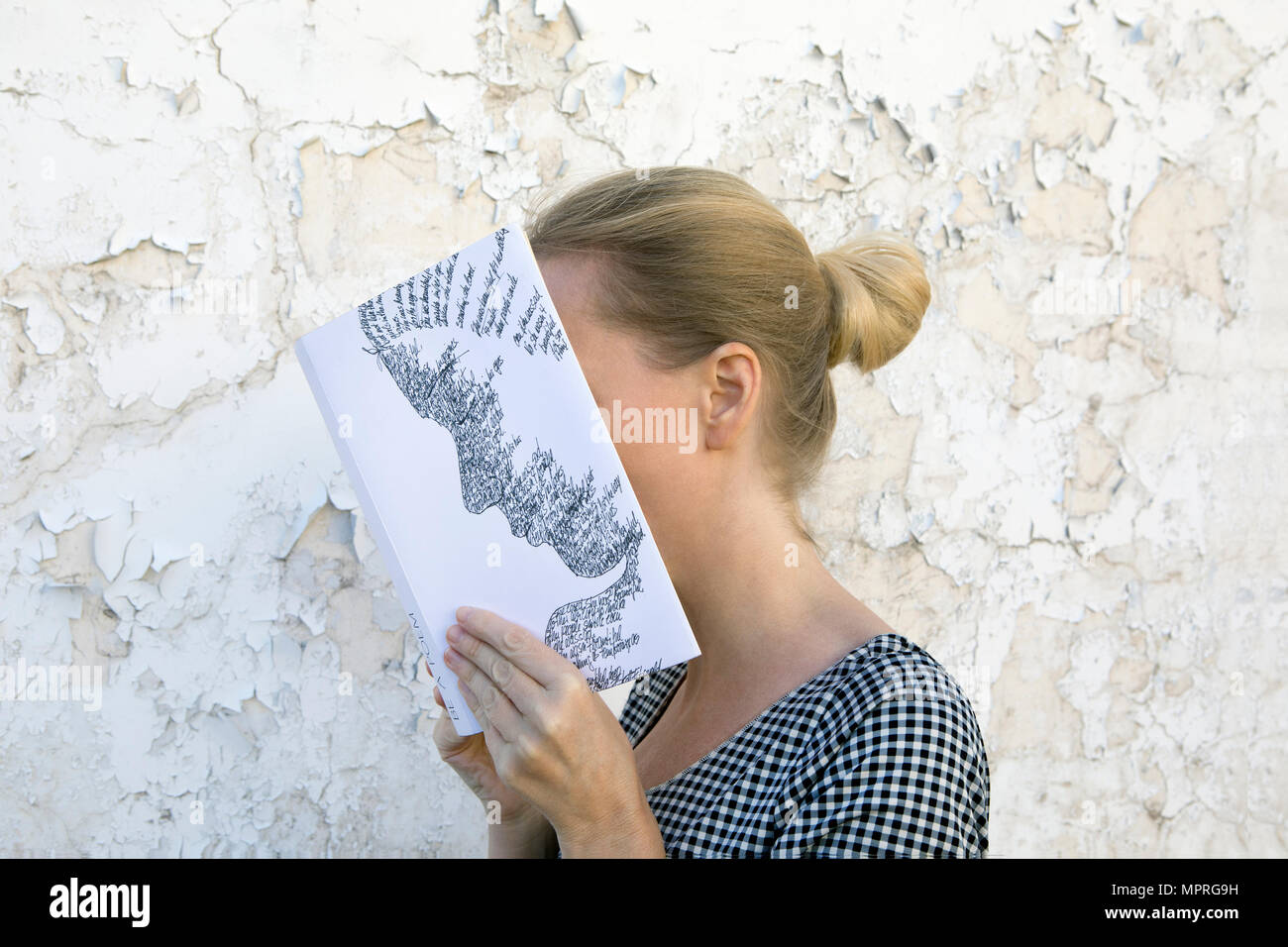 Woman covering face with book, reading poetry in front of wall Stock Photo