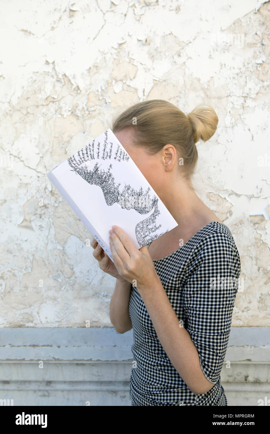Woman covering face with book, reading poetry in front of wall Stock Photo