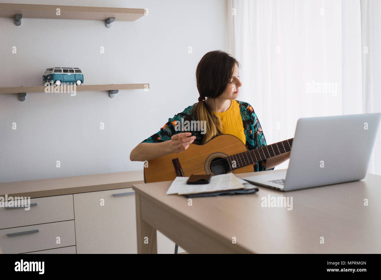 Young woman sitting at table at home with laptop playing guitar Stock Photo