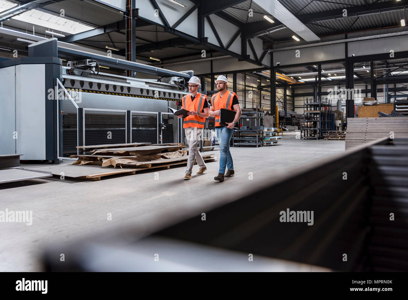 Two men wearing hard hats and safety vests walking on factory shop floor Stock Photo