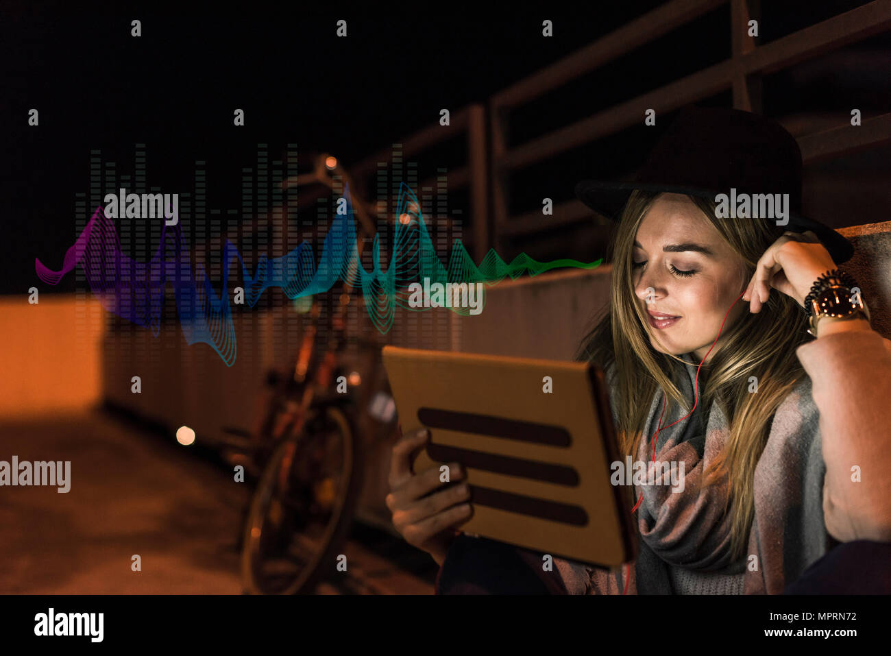 Young woman with tablet and earphones sitting outdoors at night Stock Photo
