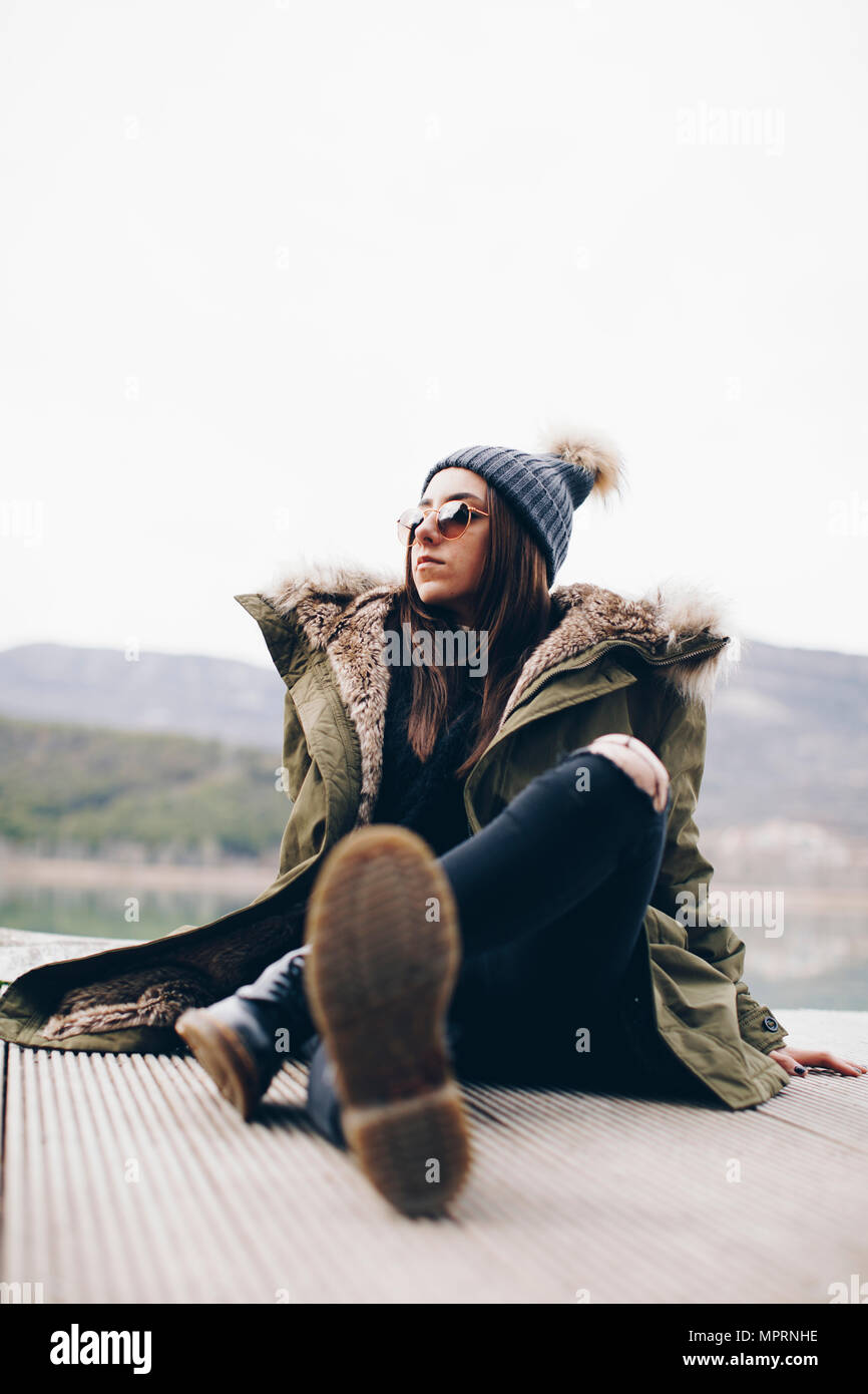 Portrait Of Young Woman Sitting On Jetty Wearing Sunglasses And