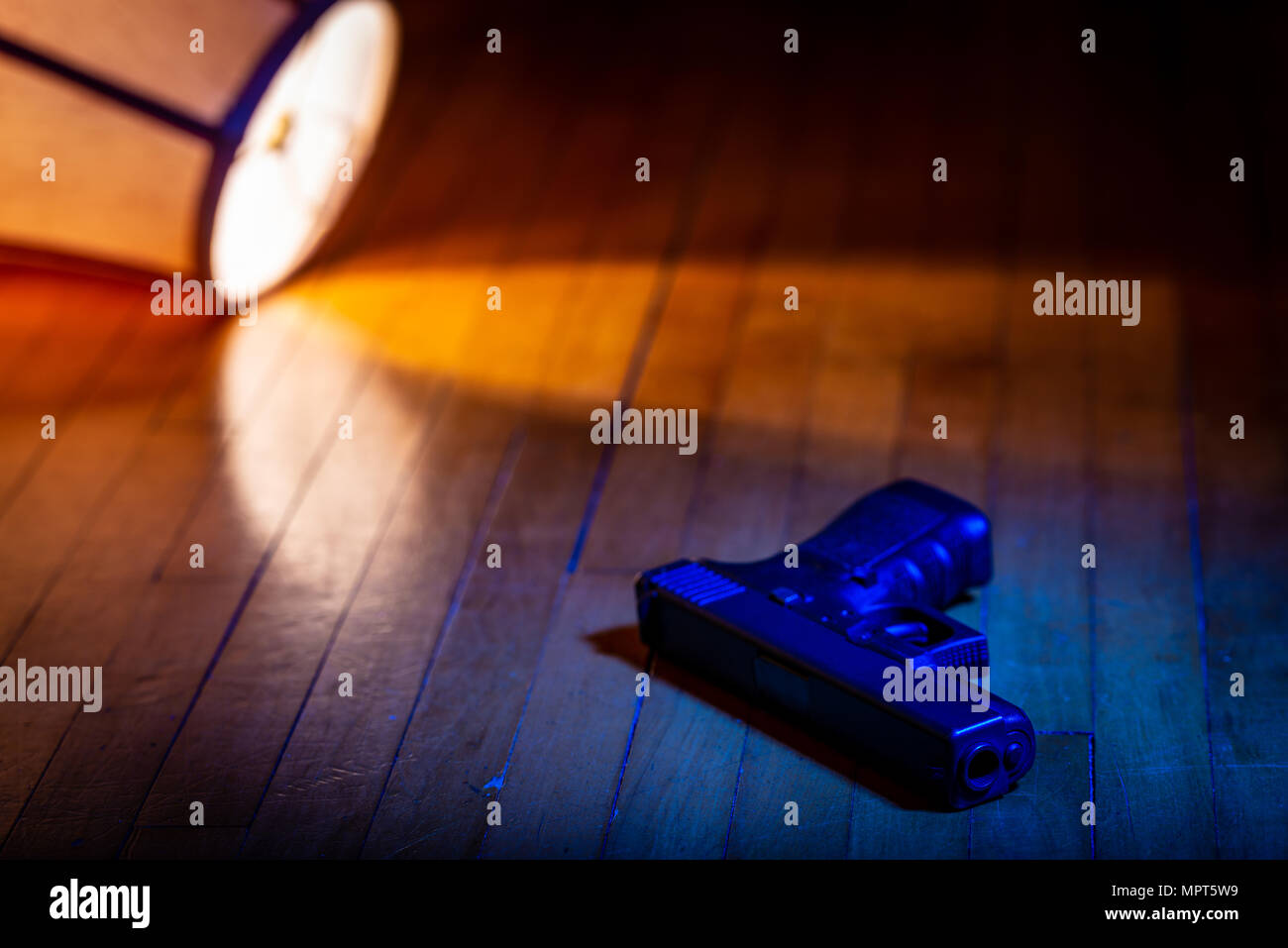 A firearm lays on the wood floor in front of a lamp which has been knocked over. Stock Photo