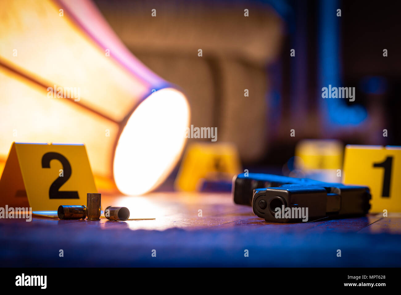A firearm and other evidence is marked with evidence markers on the floor of a home. A knocked over lamp lays nearby. Stock Photo