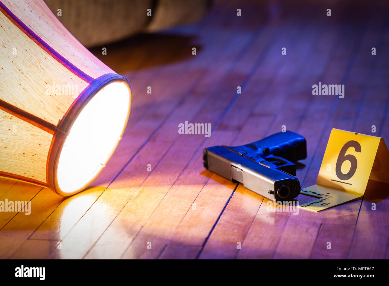 A gun is marked with an evidence marker on the floor of a home. A knocked over lamp lays nearby. Stock Photo