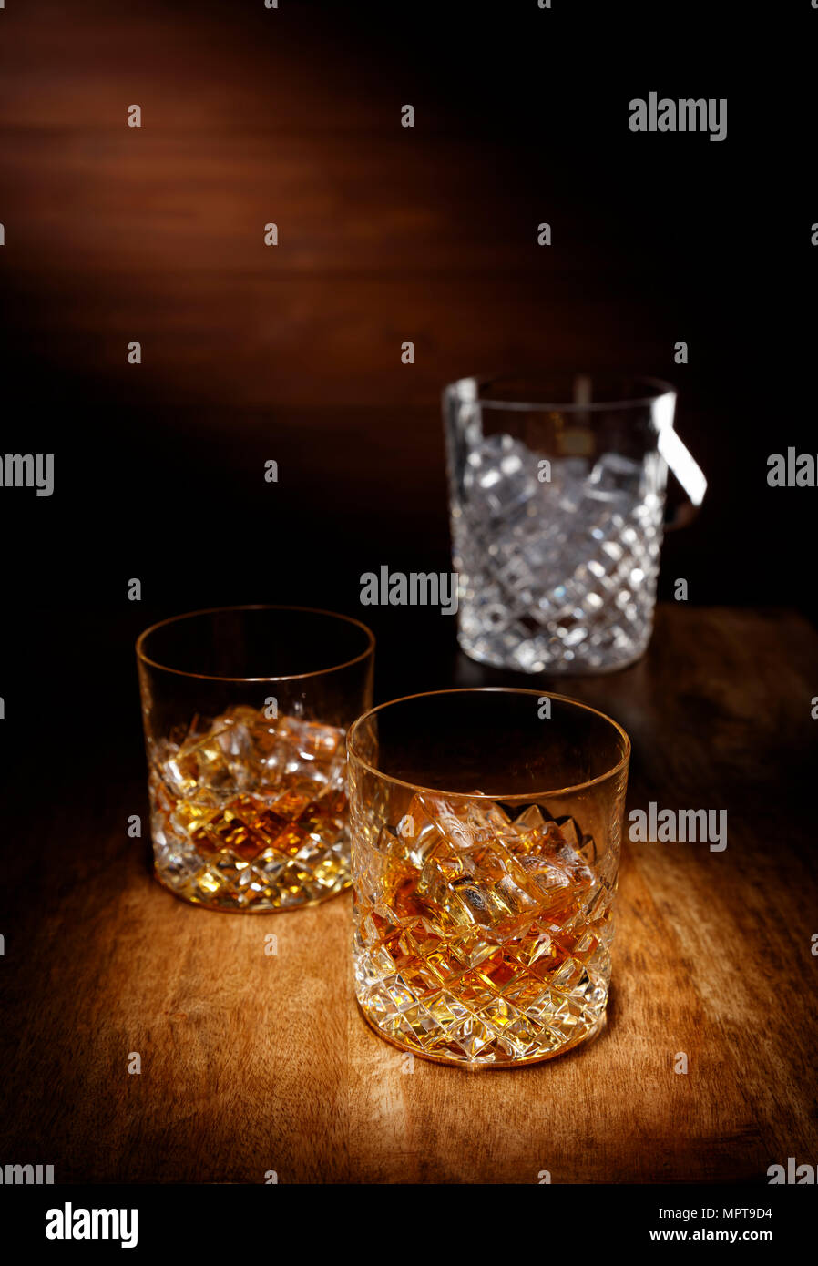 A spotlight on a two crystal glass of scotch whisky with a ice bucket in the backghround, shot on a wooden table top. Stock Photo