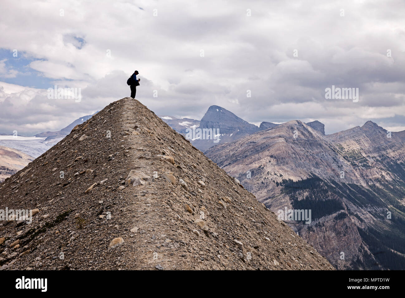 A silhouette view of an anonymous hiker stands on top of a small hill shaped like a pyramid in front of the Canadian Rockies in Yoho National Park. Stock Photo