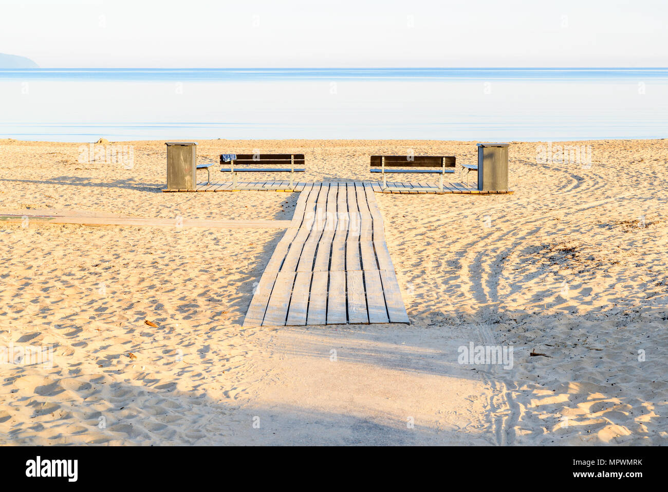 Wooden walkway at a beach, with two empty benches and trash bins on the sides. Windless sea under the horizon on a sunny morning before the bathers ar Stock Photo