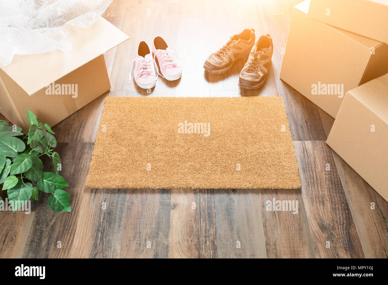 Home Sweet Home Welcome Mat, Moving Boxes, Women and Male Shoes and Plant on Hard Wood Floors. Stock Photo