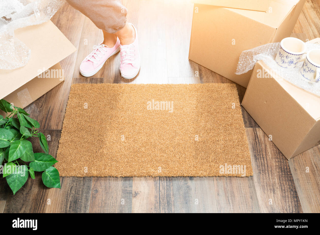 Woman in Pink Shoes and Sweats Standing Near Home Sweet Home Welcome Mat, Boxes and Plant. Stock Photo
