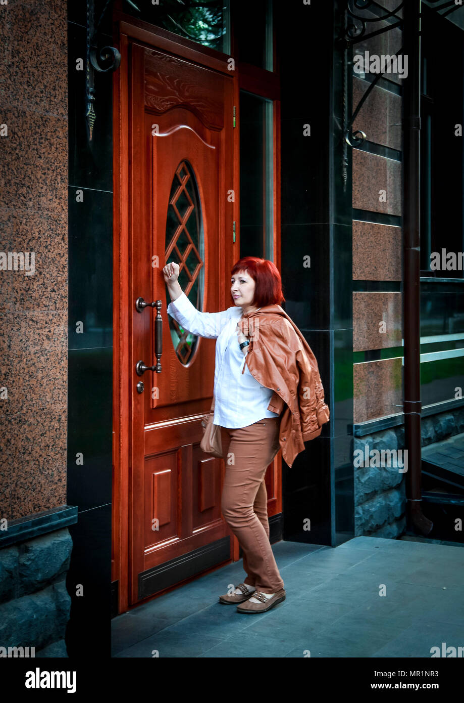 Young attractive woman with jacket on shoulder knocking on brown door Stock Photo