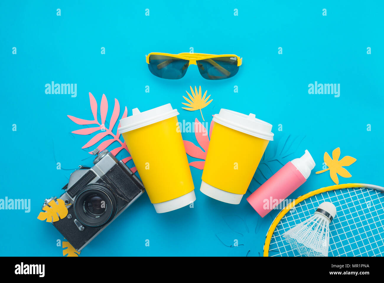 Lemonade paper cups with camera, lotion, sunglasses, badminton racket and and tropical leaves on a bright blue background. Beach and summer drink flat Stock Photo