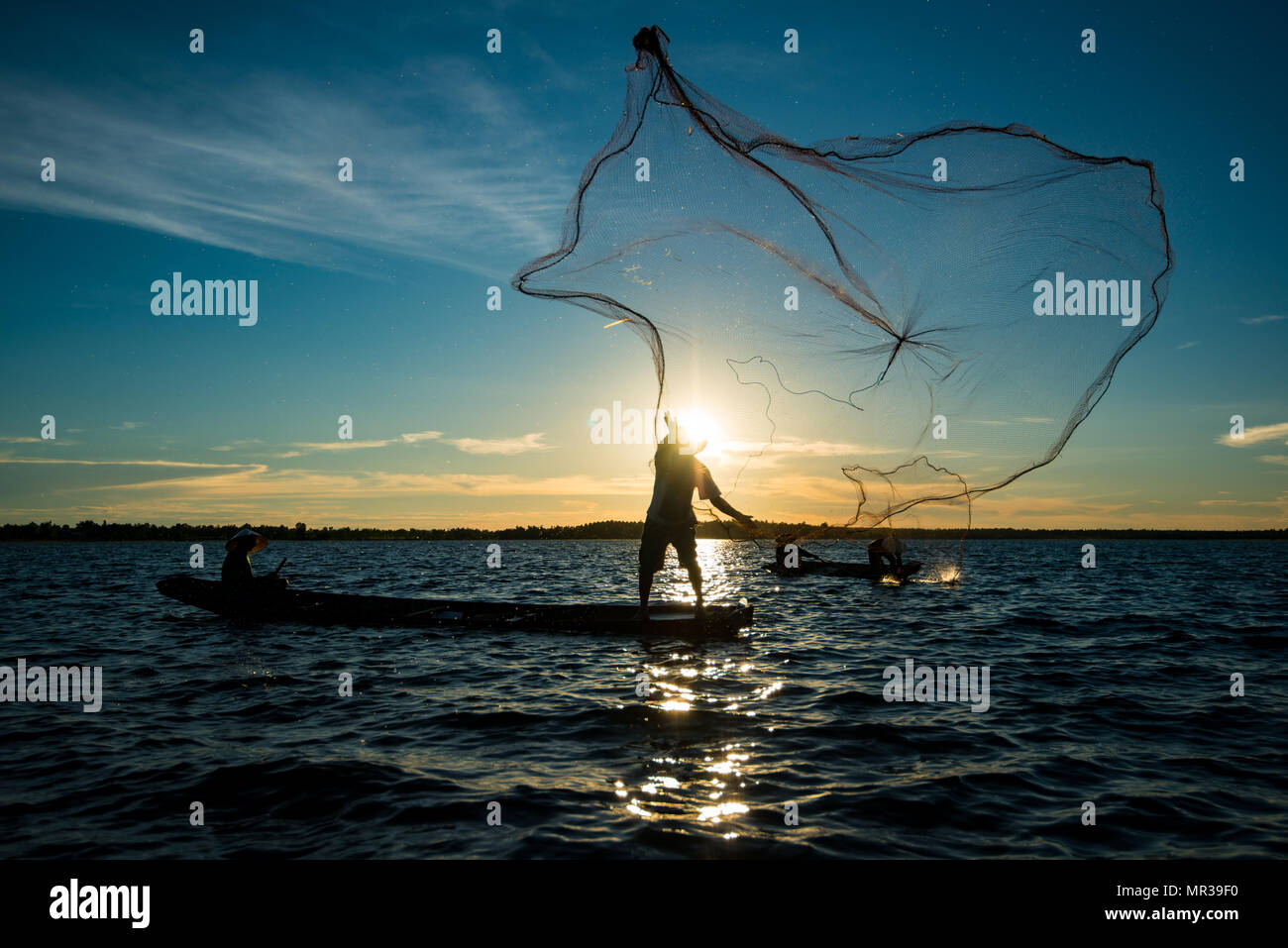 Un-identified silhouette fisher man on boat fishing by throwing fishing net to river during sunset in rural of Thailand Stock Photo