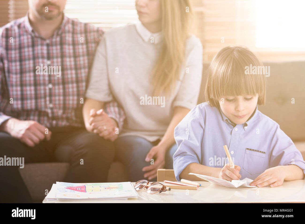 Man and woman holding hands on a couch and a boy in front drawing pictures by a table during a family psychotherapy session. Front view. Flare. Blurre Stock Photo