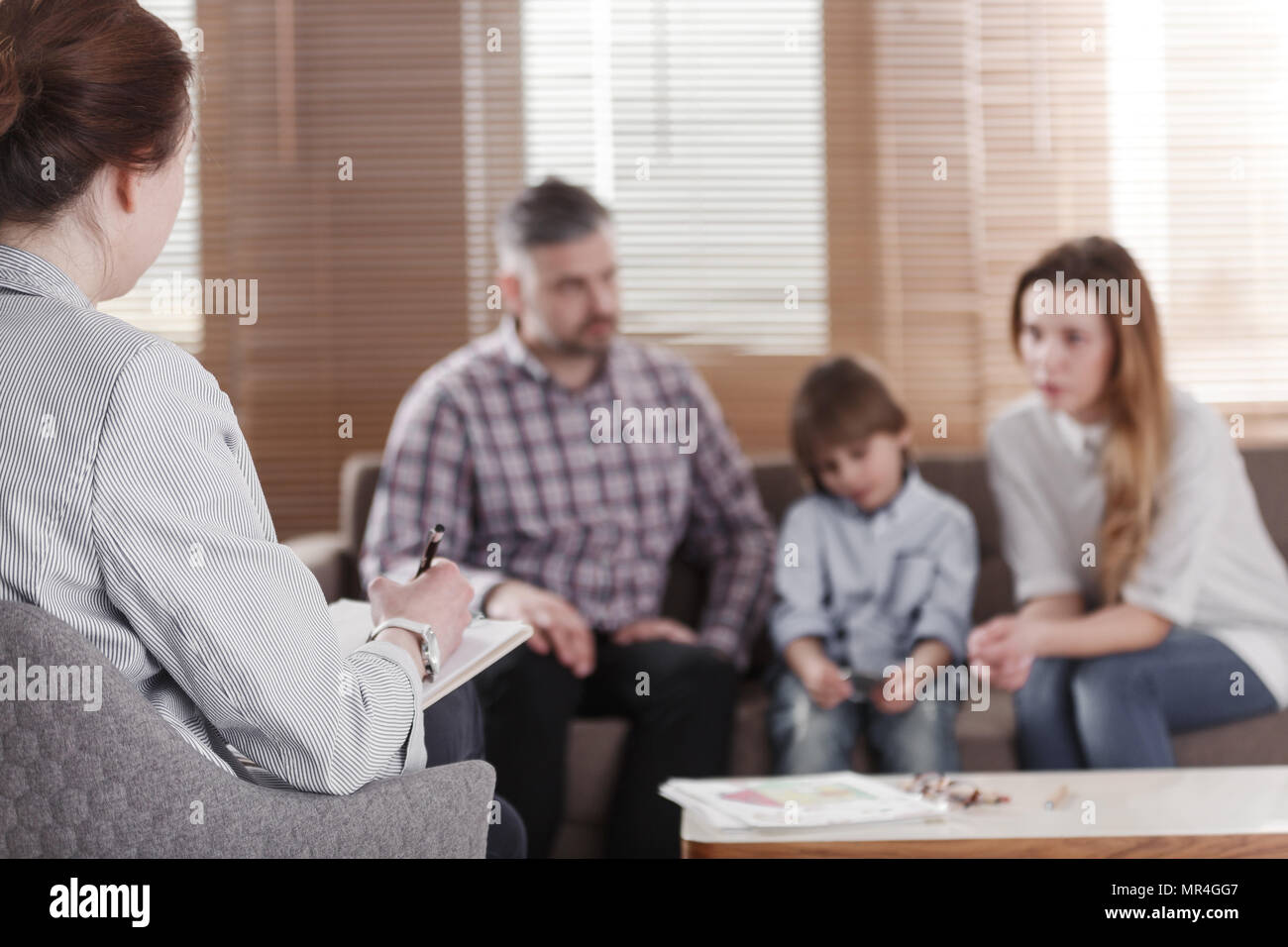Rear view of female psychologist helping young family with a kid to solve child development problems. Family sitting on a sofa in the blurred backgrou Stock Photo
