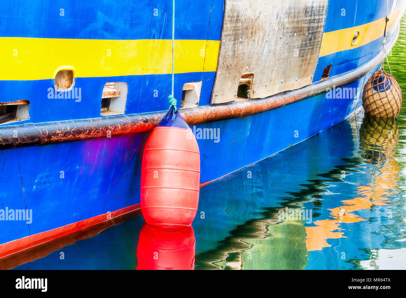 Orange and yellow bouys hang from ropes over the side of a blue and yellow boat.  Their reflections are seen in the sea water. Stock Photo