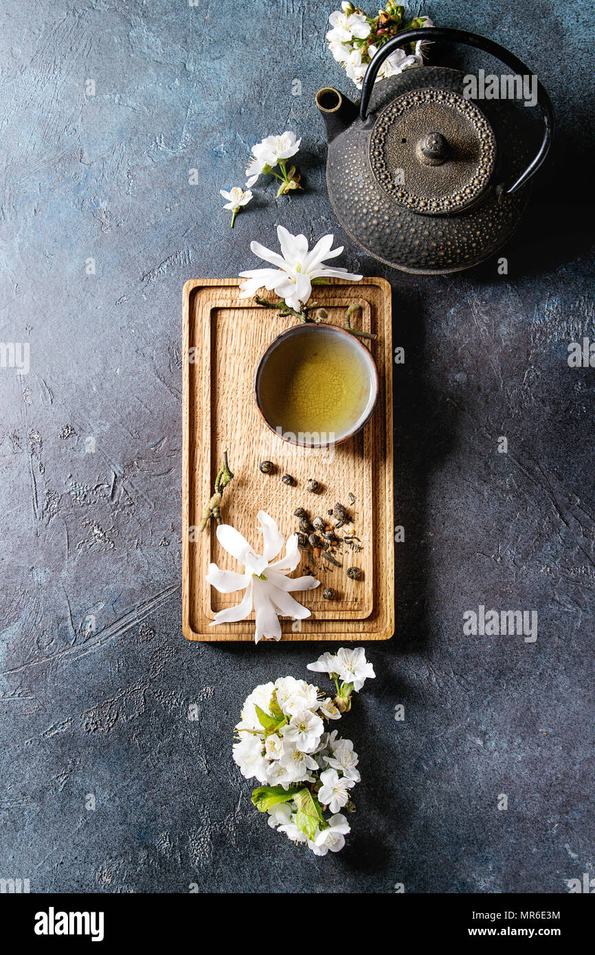 Traditional ceramic cup of hot green tea on wooden board with black iron teapot, spring flowers white magnolia and cherry blooming branches over dark Stock Photo
