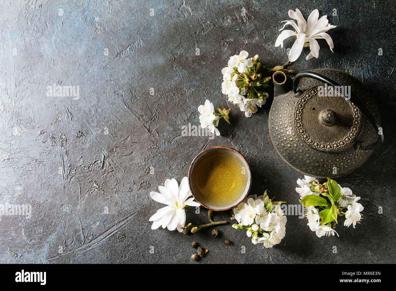 Traditional ceramic cup of hot green tea with black iron teapot, spring flowers white magnolia and cherry blooming branches over dark blue texture bac Stock Photo