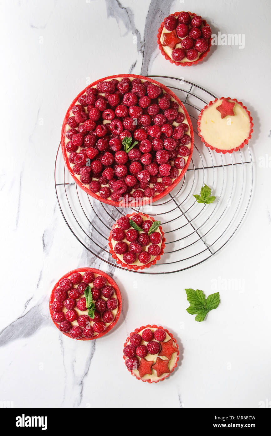 Variety of red raspberry shortbread tarts and tartlets with lemon custard and glazed fresh raspberries served on cooling rack over white marble textur Stock Photo