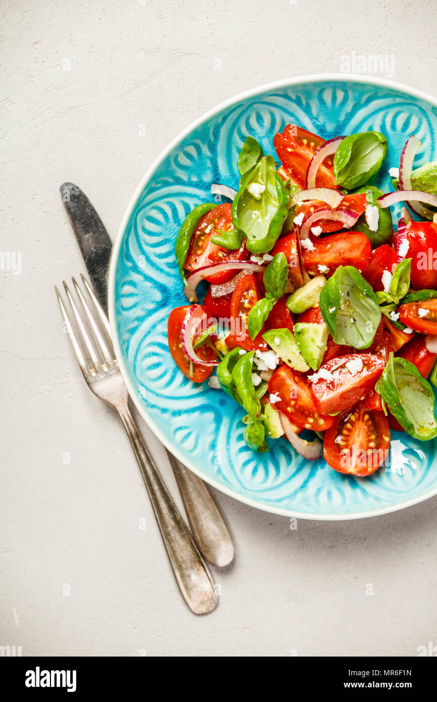 Tomato, basil, avocado and onion salad in blue plate for vegan, gluten free, allergy-friendly, clean eating or raw diet. Grey concrete background and Stock Photo