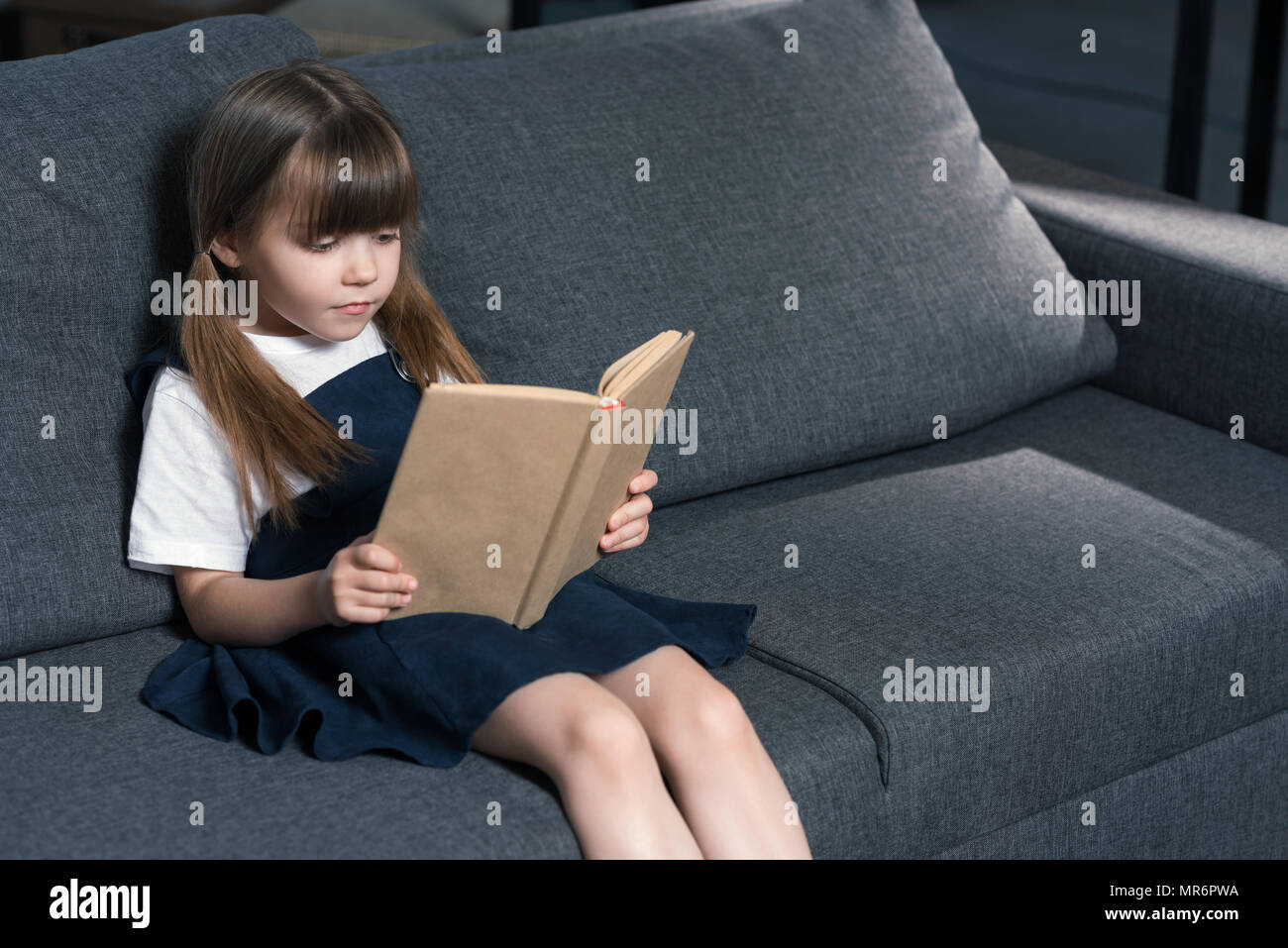 portrait of little girl sitting on sofa and reading book at home Stock ...